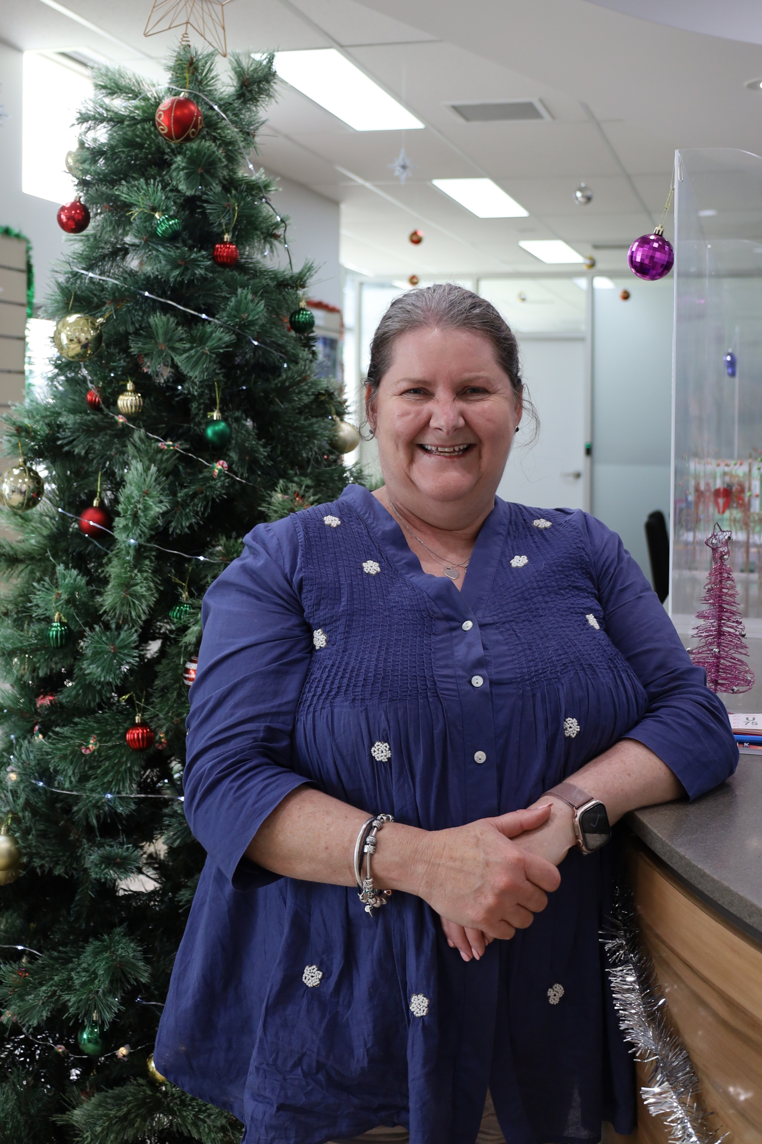 Mayor Boston stands next to a christmas tree in the Council front office. She wears a blue shirt with white embroidery and is smiling, clasping her hands in front of her.