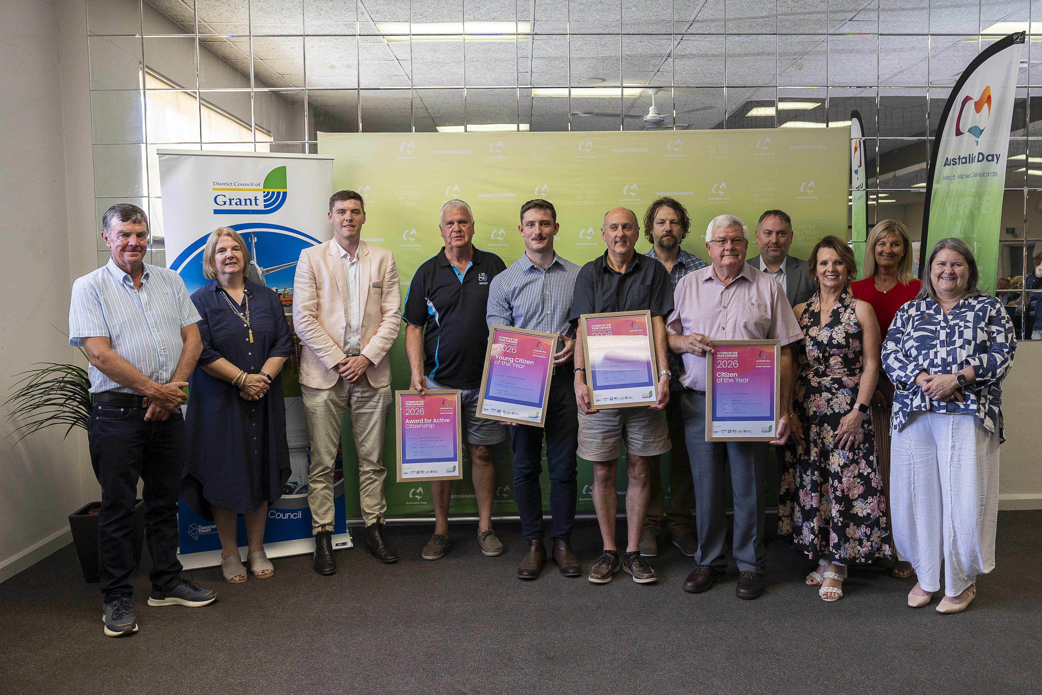A group of people stand in front of a green backdrop. Four people in the centre are holding certificates from the Citizen of the Year Awards.