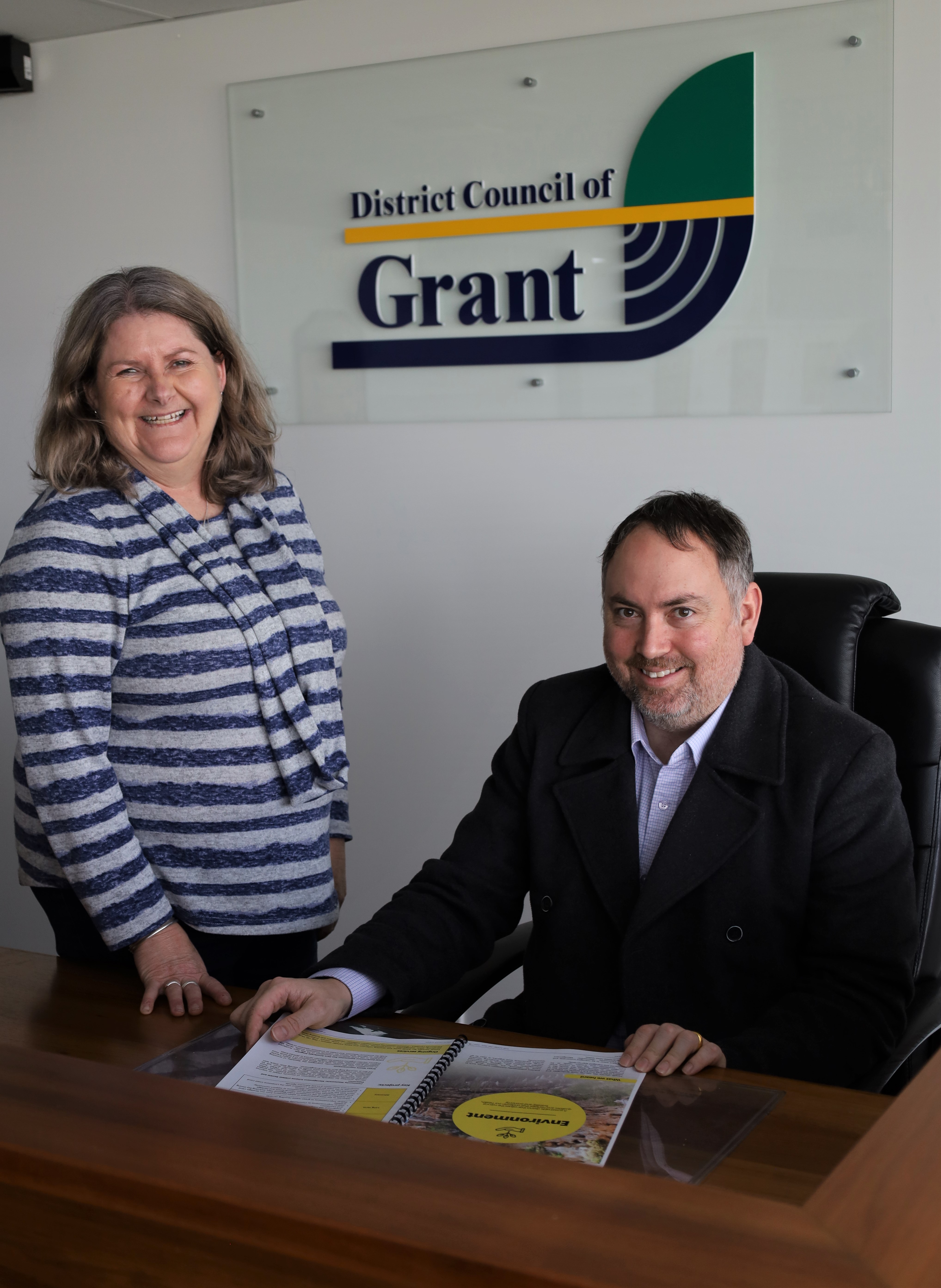 One person sitting at a desk and another leaning over the same desk smiling and reading a document